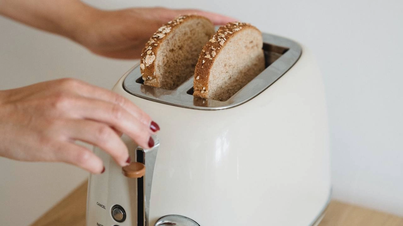 Photo. A person with red nail polish uses a white toaster to toast two slices of golden-brown multigrain bread topped with oats and seeds. Two hands grip the toaster as if about to push down the toasters' spring to start the toasting process