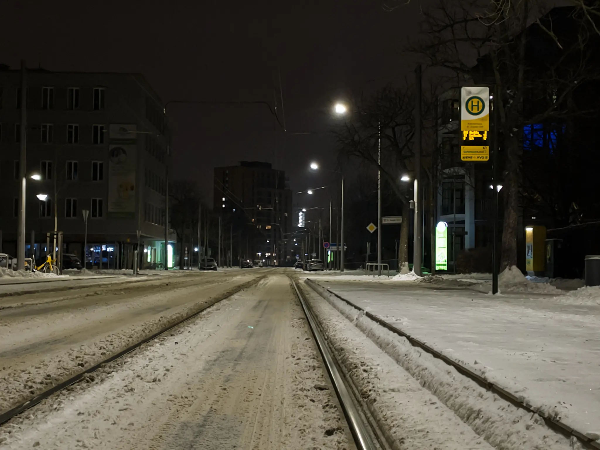 Photo of a snowy urban street at night with tram tracks running down the center. The street is covered in white snow and ice, with clean metal rails embedded in the snow. On both sides are multi-story buildings with lit windows, street lamps casting yellow light, and bare trees. On the right side is a yellow tram stop sign with a green-on-yellowH symbol. The sky is dark gray, and the scene is illuminated by various street lights creating a dim, lonely snow nighttime atmosphere.