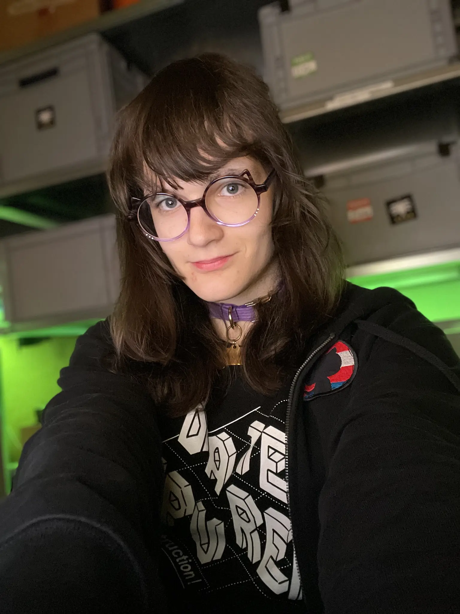 Photo. Person with long brown hair and purple round glasses tilts their head slightly while looking at the camera with a subtle smile. They wear a black hooded jacket with a transgender colored github logo patch on it, a purple collar with a gold bone charm, and a black shirt with white 3D text