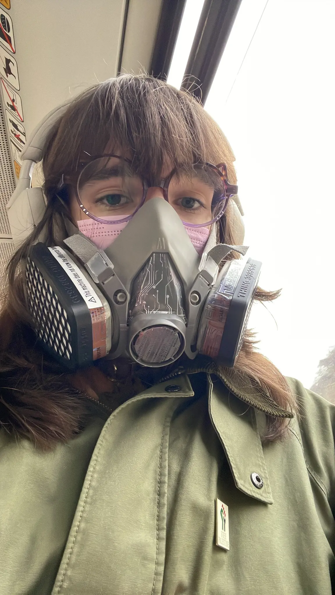 Photo of Ava sitting in a public transport vehicle. It is wearing an olive green jacket with a Free Palestine pin on it. Its brown hair is shoulder length. It is wearing a pink surgical mask underneath their respirator, which is decorated with PCB-like trace designs. Ava is also wearing its signature purple-brown glasses with little cat ears on them.