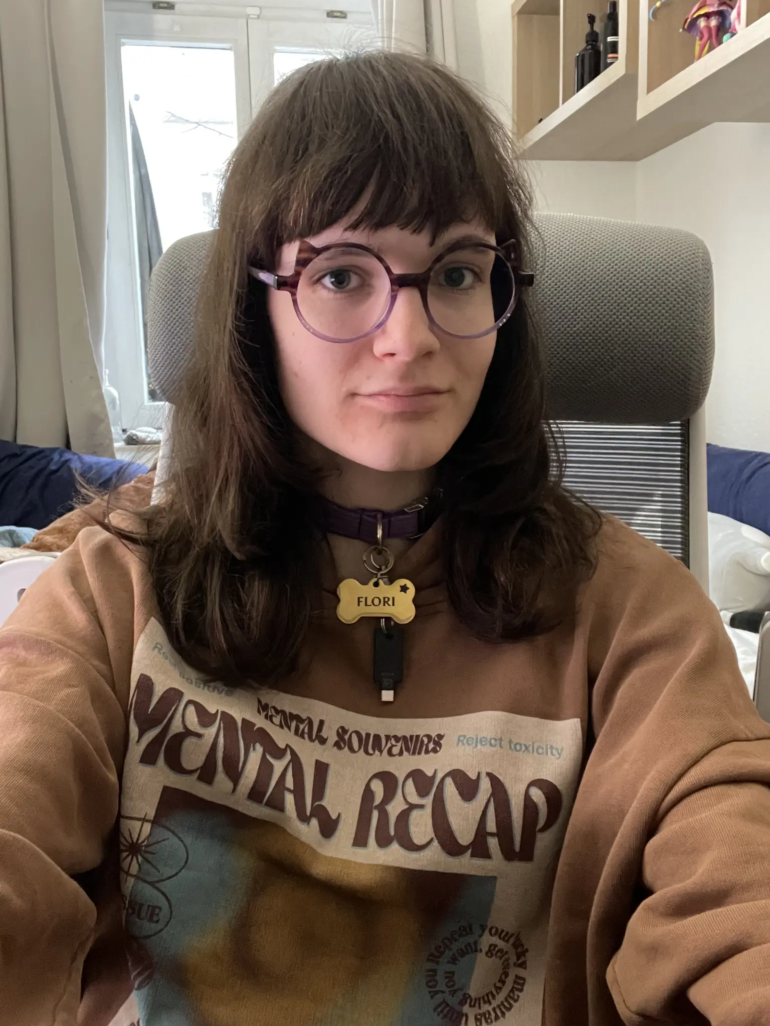 Photo of ava, a person with shoulder-length brown hair wearing purple-framed round glasses and a tan/brown print hoodie with text reading MENTAL SOUVENIRS, Reject toxicity, and MENTAL RECAP. They are wearing a purple collar with a bone-shaped golden tag that reads FLORI, and a black USB-C YubiKey dangling from that collar. The setting is an indoor living space with a gray textured couch or chair behind them, white cabinets and shelving visible in the background, and natural light coming from a window.