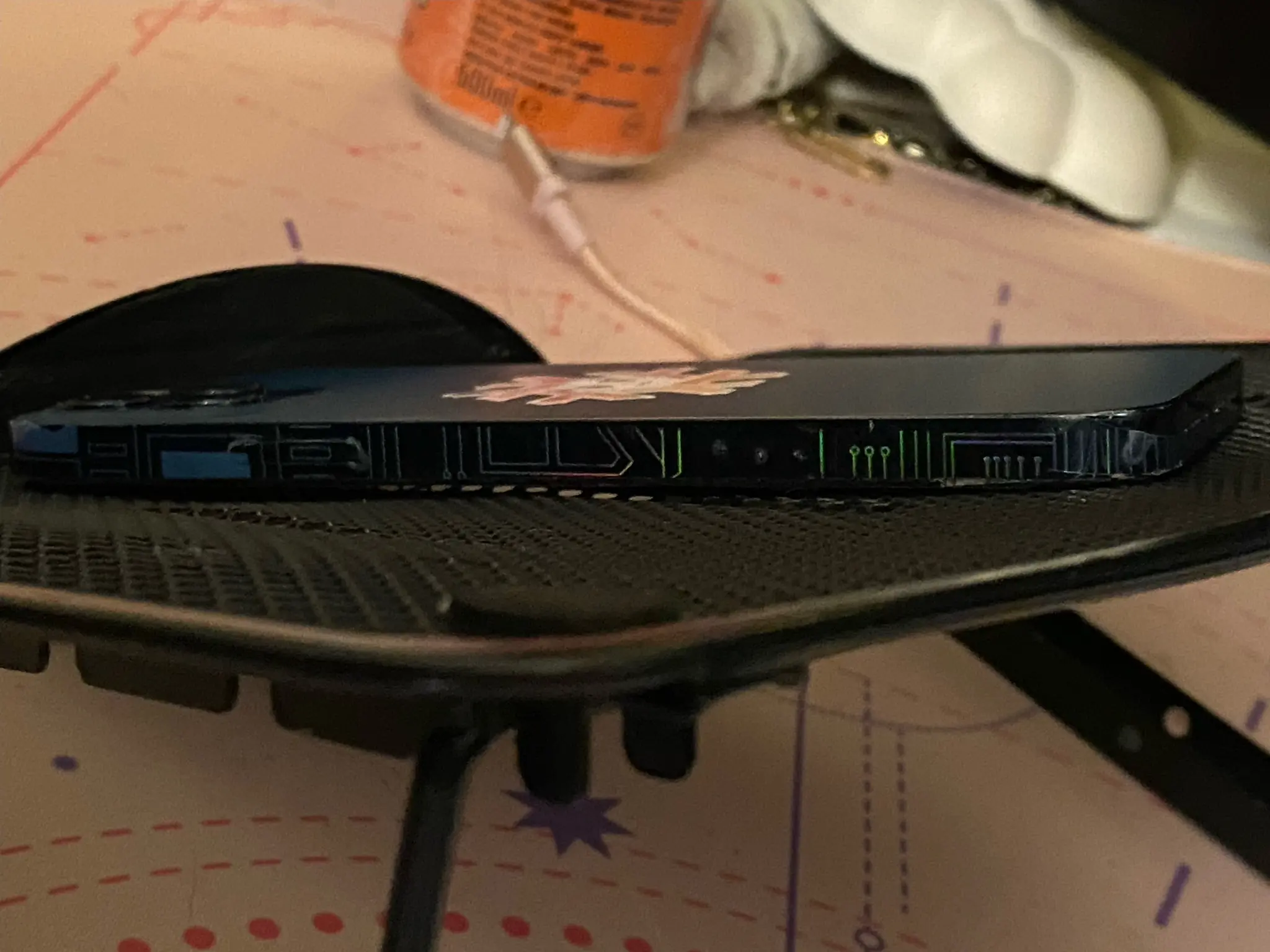 A dark-blue iPhone 12 Pro propped up on a tablet stand. Photograph taken from the side. On the flat sides of the device, traces and PCB-like structures can be seen, as if printed on. These structures seem to illuminate in different colors when hit by light in a certain way. With red, orange, yellow and green hues visible shining on the otherwise gray or light blue PCB traces.