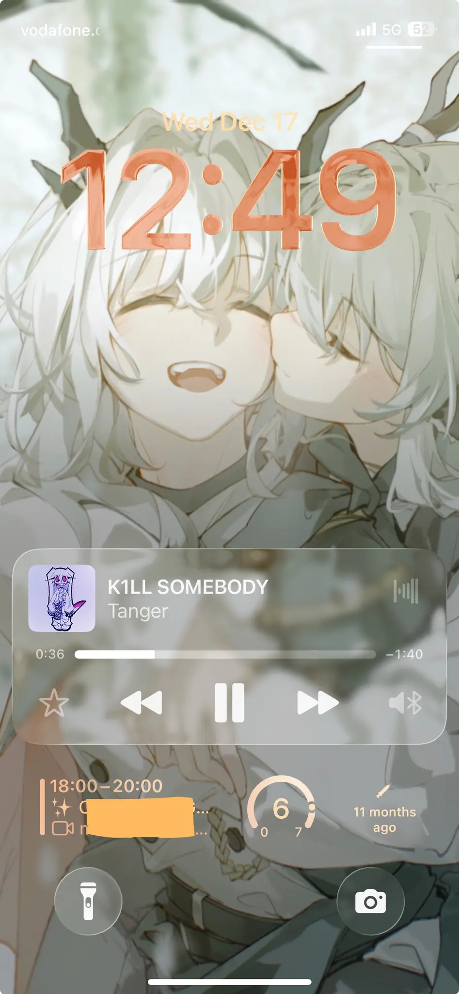 Photo of a smartphone lock screen displaying the time 12:49 in large orange numbers at the center. The background features anime-style artwork of a character with light blonde or white hair and horn-like protrusions, surrounded by green and sage-colored leaves. At the top of the screen, it shows “vodafone.c” and “5G” with signal indicators. A semi-transparent music player widget is visible in the middle, showing the song “KILL SOMEBODY” by “Tanger” with a small purple character icon, playback controls including previous, pause, and next buttons, and a progress bar showing 0:36 of 1:40. Below the music player is an orange rectangular element with text “18:00-20:00” and “11 months ago” with a circular indicator showing “6”. At the bottom are two circular buttons: a flashlight icon on the left and a camera icon on the right.