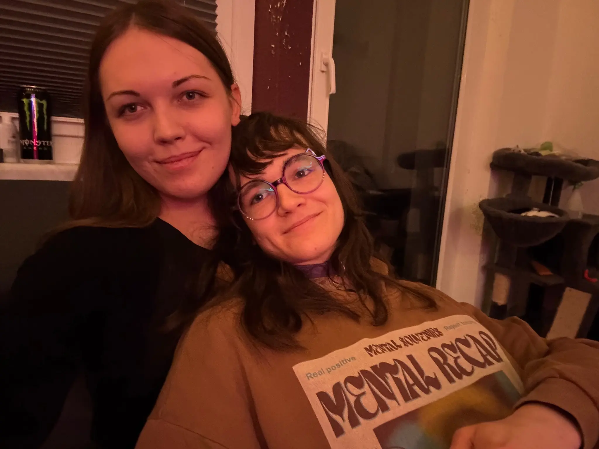 Photo of two feminine people taking a selfie together indoors. The woman on the left has long, straight dark brown hair, defined eyebrows, and wears a black top. The person on the right has wavy, shoulder-length dark brown hair with lighter highlights, wears large round purple-framed glasses with cat ears on the frame. Both are cuddling with each other and looking at the camera, smiling.