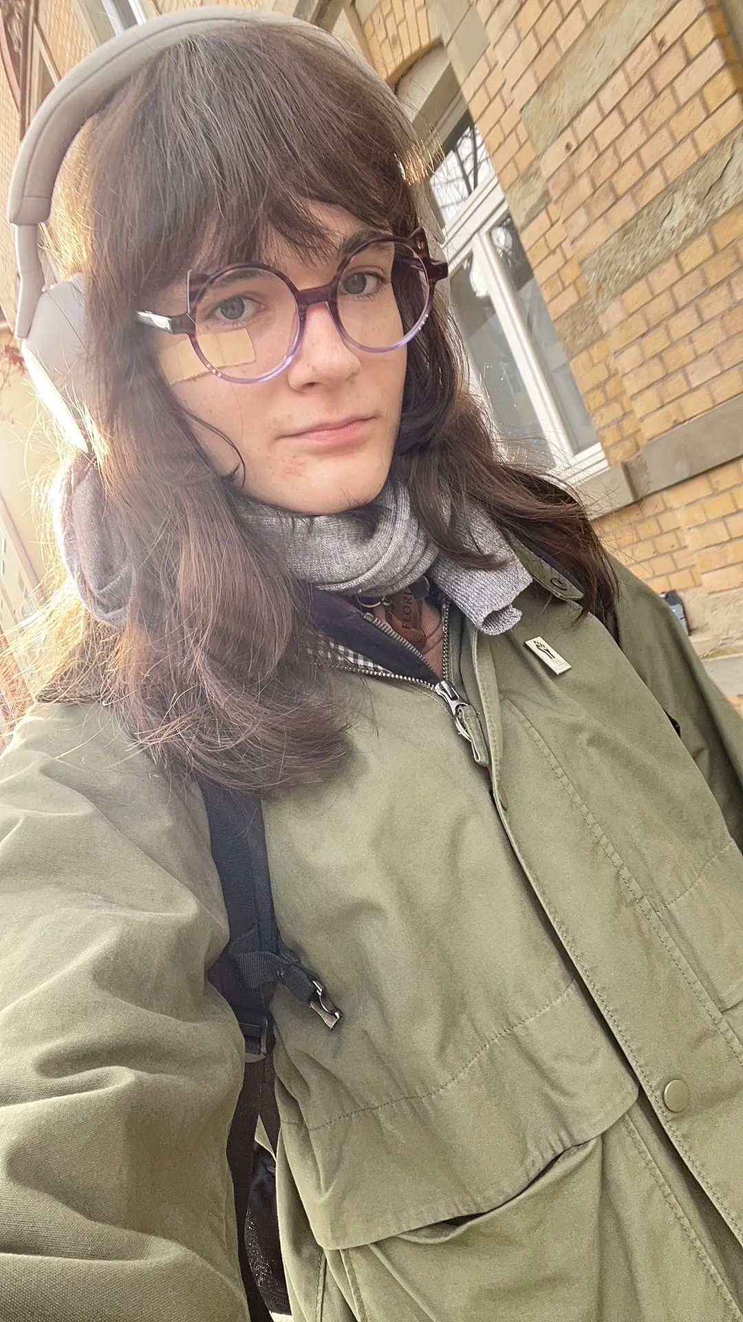 Photo of a person taking a selfie outdoors in front of a yellow brick building with white-framed windows. The person has shoulder-length brown hair, wears large purple-framed glasses, white over-ear headphones, a gray knit scarf, and an olive green jacket. The lighting is warm and wintery