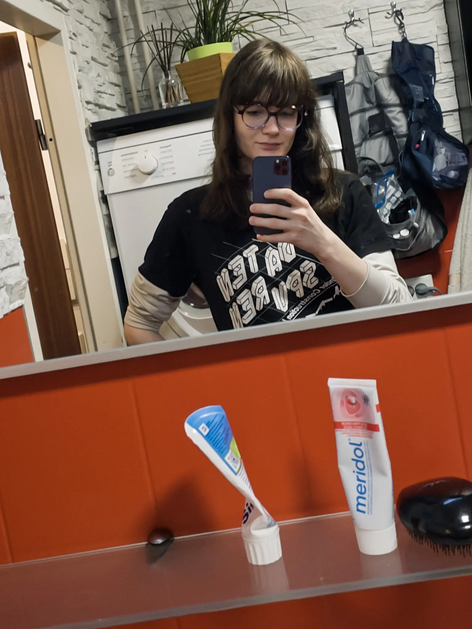 Photo of a person taking a mirror selfie in a bathroom. They have shoulder-length dark hair, wear glasses, and a black graphic t-shirt with beige long sleeves underneath. They hold a dark phone in front of a mirror with white brick-pattern wallpaper behind them. The orange bathroom counter in the foreground displays a Meridol toothpaste tube, a toothbrush in a holder, and a black hairbrush. A backpack hangs on the right side behind the person, and a white washing machine with a potted plant on top sits in the background.