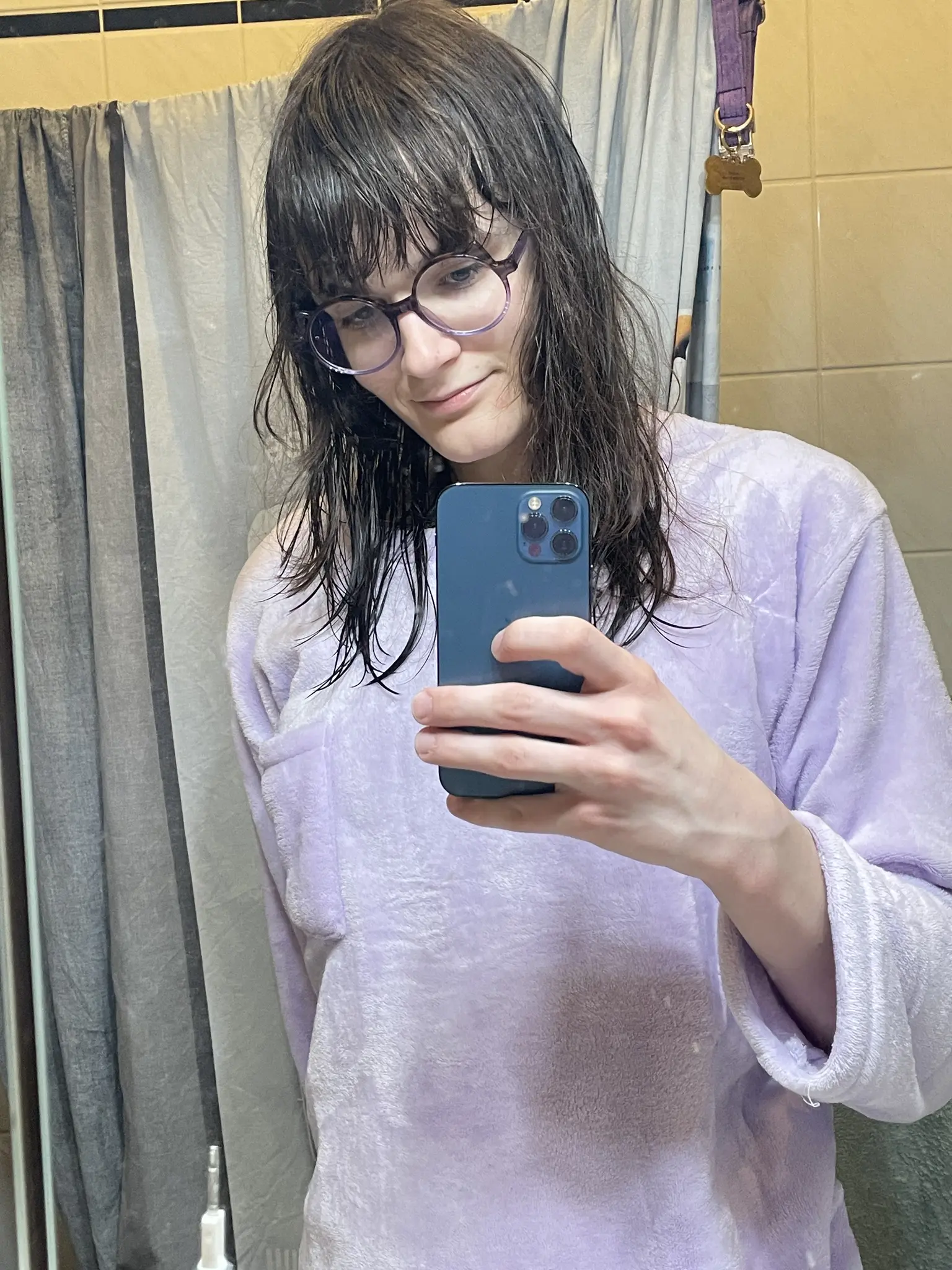 Photo of a person taking a mirror selfie in a bathroom, wearing a light purple, fluffy oversized pajama top and large round glasses with dark frames. The person has wet, dark hair going just over the shoulders, with the hair in the back longer than the hair in the front and is holding a dark blue iPhone. Behind them is a clothes line with a purple dog collar hanging on the right side and white tile walls which look yellow in the warm white light