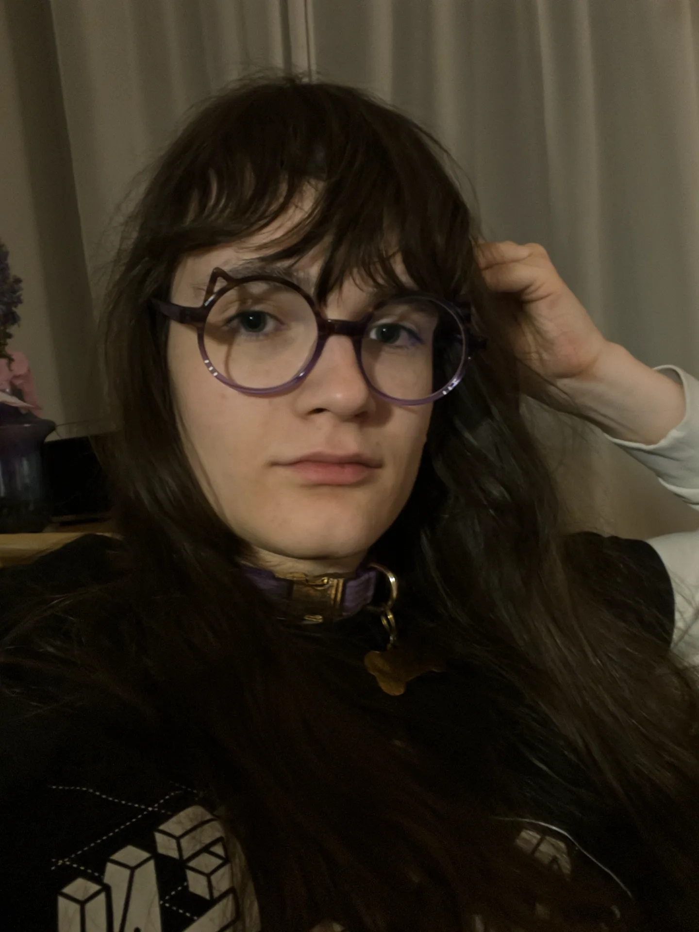 Photo of a person with shoulder-length brown hair and large purple round glasses with little cat ears looking directly at the camera with a neutral, satisfied expression. They are raising their right hand to their hair near their ear. The person is wearing a black shirt with isometric font pattern design visible at the bottom. Around their neck is a purple collar with gold-colored hardware and a gold bone-shaped tag. Behind them is a beige curtain, and partially visible on the left side is a dark green vase on a wooden table.