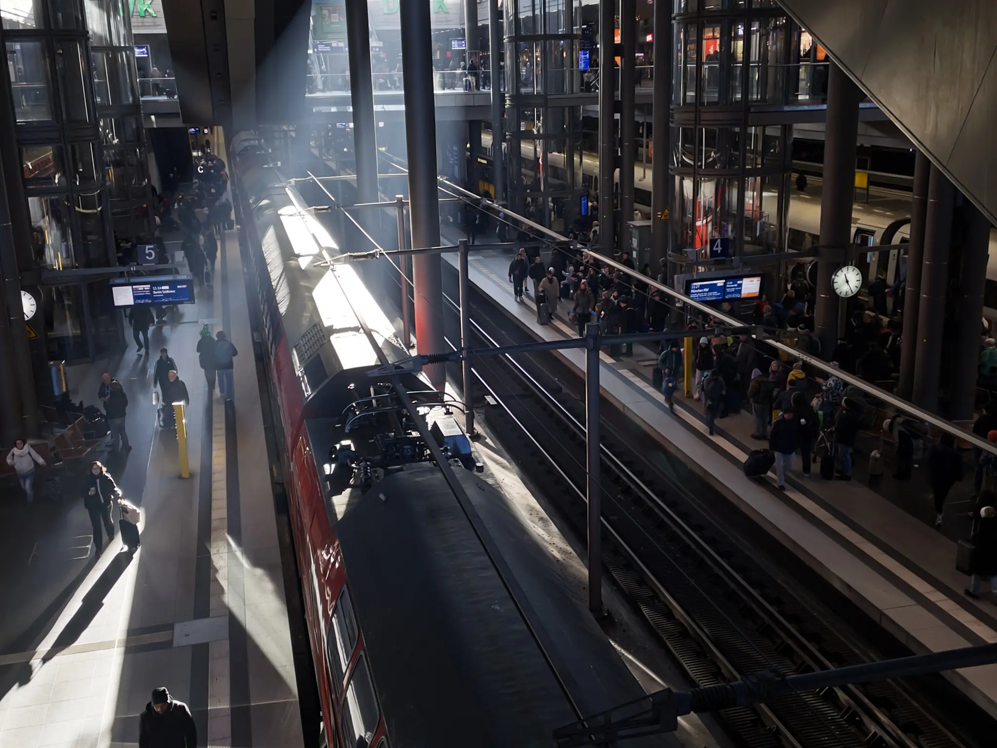 Photo of a modern train station interior taken from an elevated perspective. A red and black passenger train is stopped on the left platform, crowds of people wait on the right platform. The station features industrial, perhaps brutalist architecture with black metal framework, glass walls, and exposed concrete. Bright natural sunlight streams through the picturw, creating sharp geometric shadows and a sharp, bright streak of light across the gray platforms and floors. The lighting makes for strong contrast between the illuminated areas and darker sections. Multiple levels are visible with stairs and railings, digital information displays showing blue screens and information about departing trains, and a round clock mounted on the right side