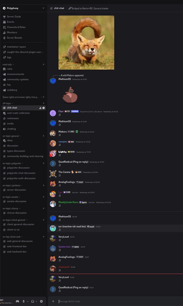 Screenshot of a Discord chat interface in the Polyphony Guild showing the chit-chat channel. The chat shows a conversation with multiple users including Mathium05, Flori, Makuru, Campos, Lighty, QuadRadical, The Canine, AnalogFeelings, MaddyUnderStars, ezr, VeryLoud, Quaternion, and chaoswolf posting messages. The first message reads "A Wild Makuru appeared" followed by an image of a red fox crouching playfully on dry grass with bright orange-red fur, white facial markings, pointed ears, and a bushy tail against a blurred natural background. The other users all posted messages in reaction to this message, each user replying with ":3" for a total of 18 messages responding with ":3".