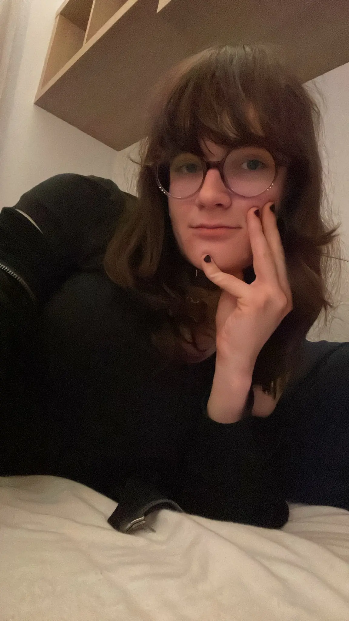 Photograph. Ava is laying on her side in a bed, looking at the camera, smiling, one hand with dark nail polish holding her chin. A bit of her lower arms are uncovered from the sweatjacket it is wearing