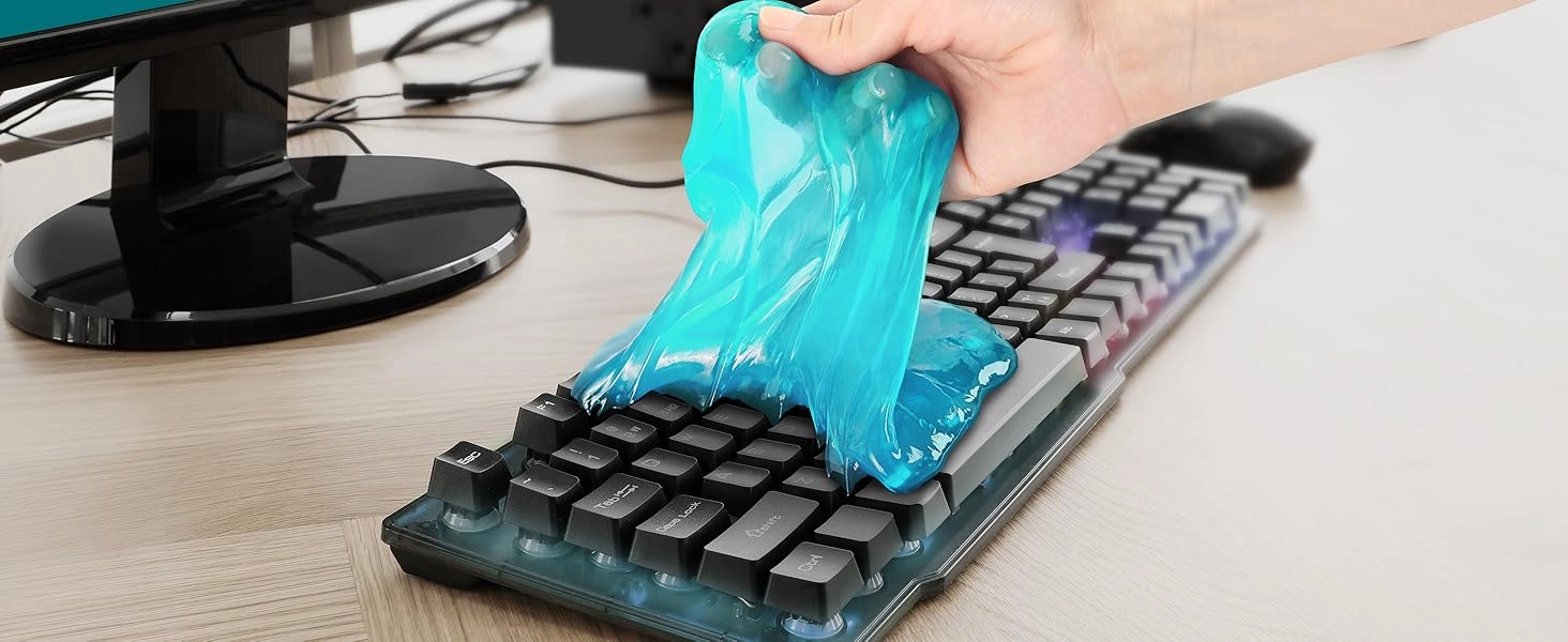 A close-up photo displays a hand pressing a mass of translucent bright blue gel onto a black mechanical keyboard resting on a light wood-grain desk. The blue slime stretches from the person's fingers and spreads across the keys on the left side, filling the crevices between buttons labeled Tab, Caps Lock, Shift, and Ctrl. A black monitor stand and a computer mouse are blurred in the background.