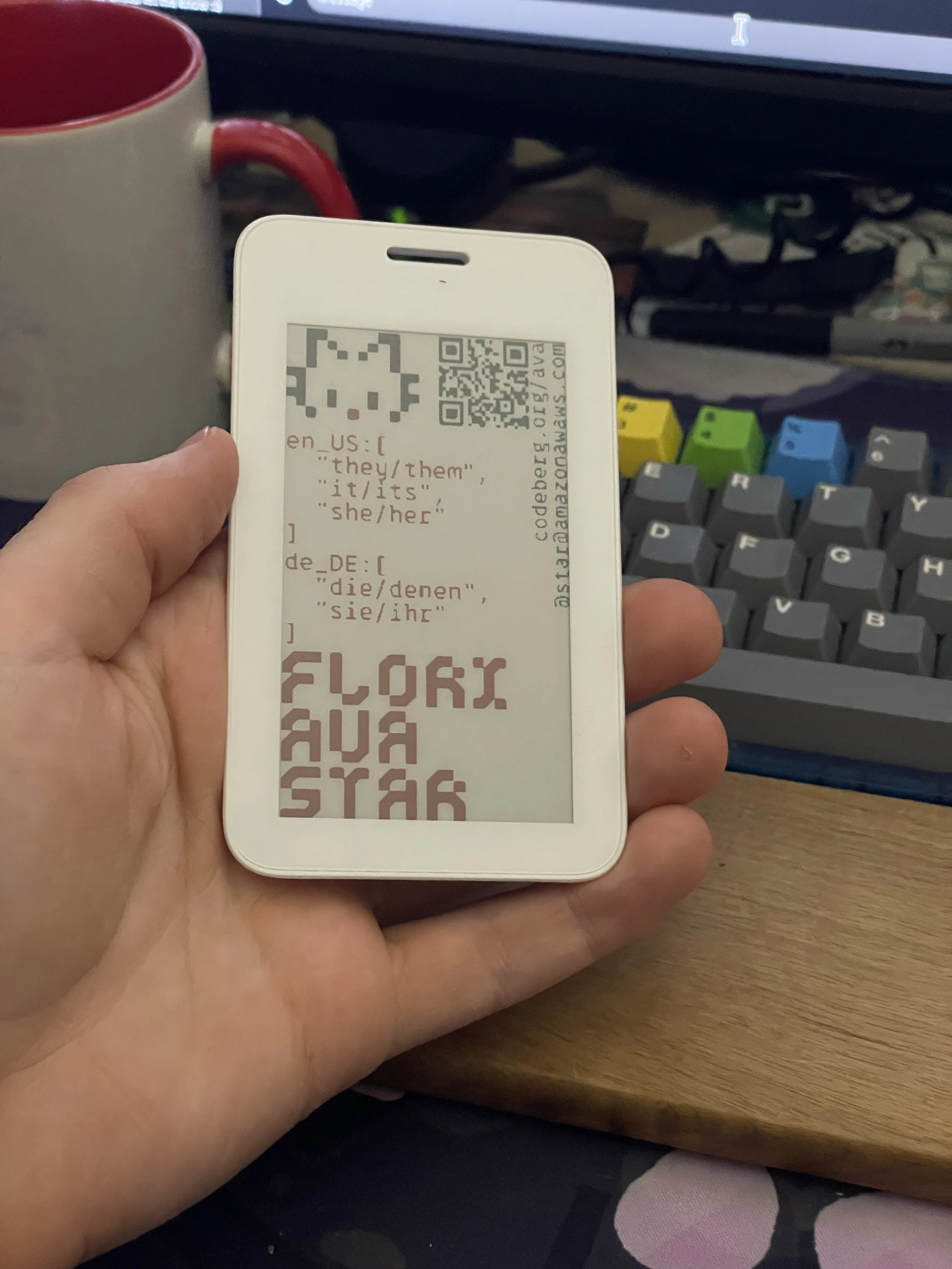 Photograph of a hand holding an e-ink badge with black, red and white colors on the display. The 9:16 aspect ratio screen displays a pixel art head of a cat in the top left corner, followed by a qr code leading to https://codeberg.org/ava in the top right corner. below these two items, a JSON object with two items is printed in red color, reading:
en_US: [
  "they/them",
  "it/its",
  "she/her"
]
de_DE: [
  "die/denen",
  "sie/ihr"
]
Below this array, the words
FLORI
AVA
STAR
are written in red, cyberpunky font with one word one each newline. On the right side, rotated by 90 degrees, black text reads "codeberg.org/ava" on the first line, and "@star@amazonawaws.com" on the second line.