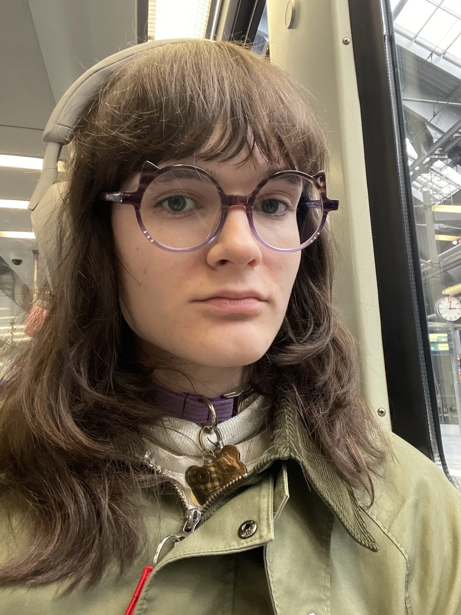 selfie of an ava with medium-length dark brown hair and bangs, wearing round purple  glasses with cat ears on them, beige/white over-ear headphones, a purple collar with a gold charm pendant, and an olive green jacket. The person is inside a public transport vehicle, and looking at the camera with a neutral, slightly content expression. Train station with a clock visible through the window behind them.