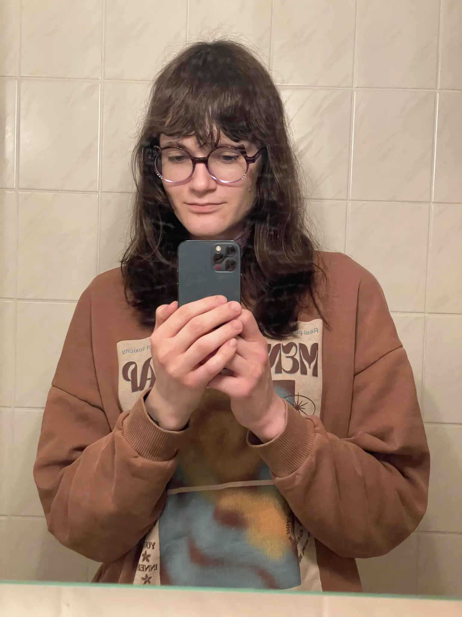 Photo of a person with dark brown shoulder-length wavy hair and purple-framed round glasses taking a mirror selfie with a dark blue smartphone. The person is wearing a brown oversized hoodie with graphic text and images printed on it. The background shows white tiled walls with a subtle diagonal pattern. The person is looking at the phone while taking the picture