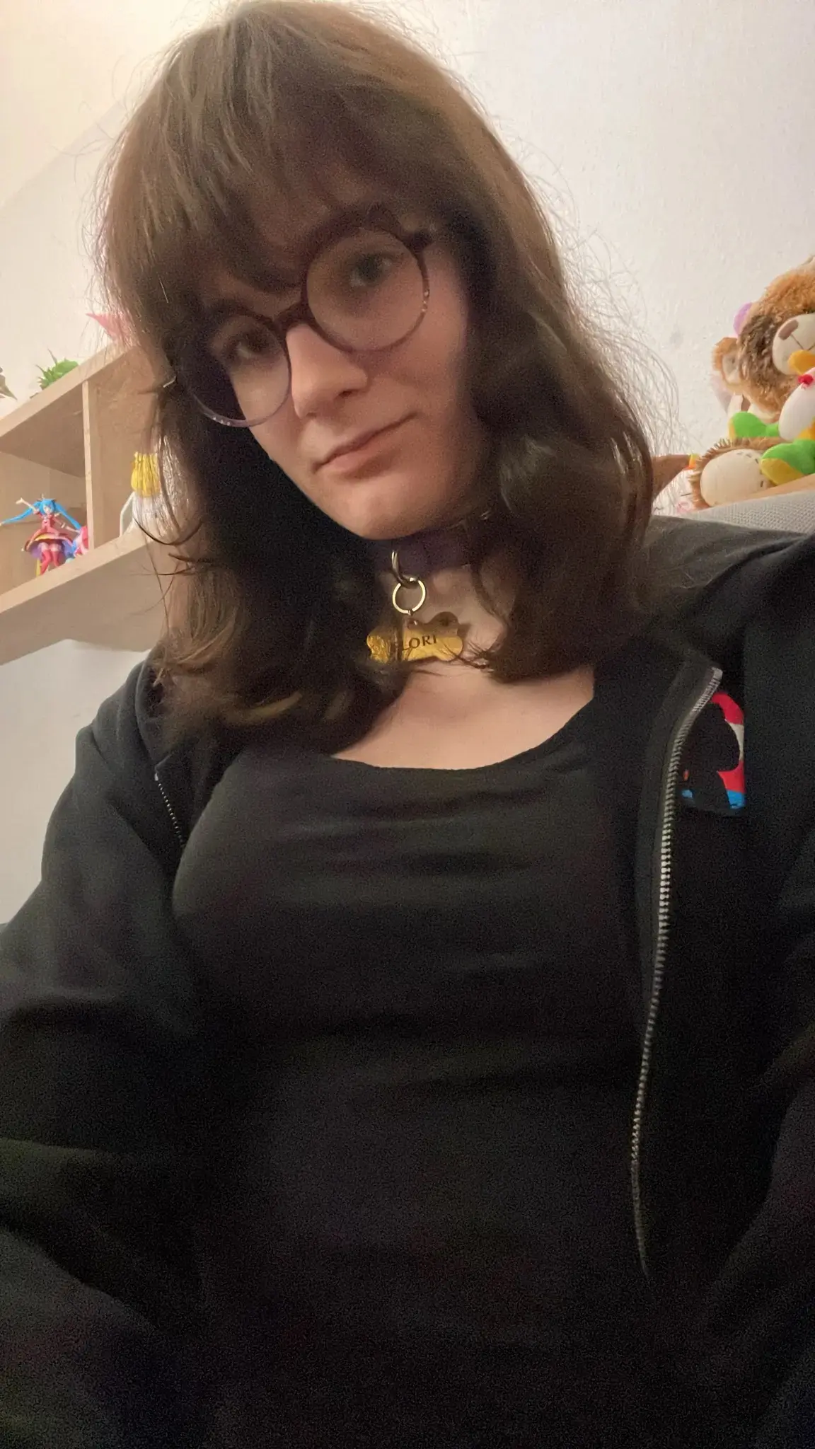 a picture of ava. Ava has collarbone-length straight brown hair and is wearing a purple collar with a golden nametag reading "Flori" on it, a black tshirt with cleavage and a black sweatjacket with a transgender themed github icon on it, zipped open. Ava is looking at the camera and smiling. Avas boobs look large in this picture