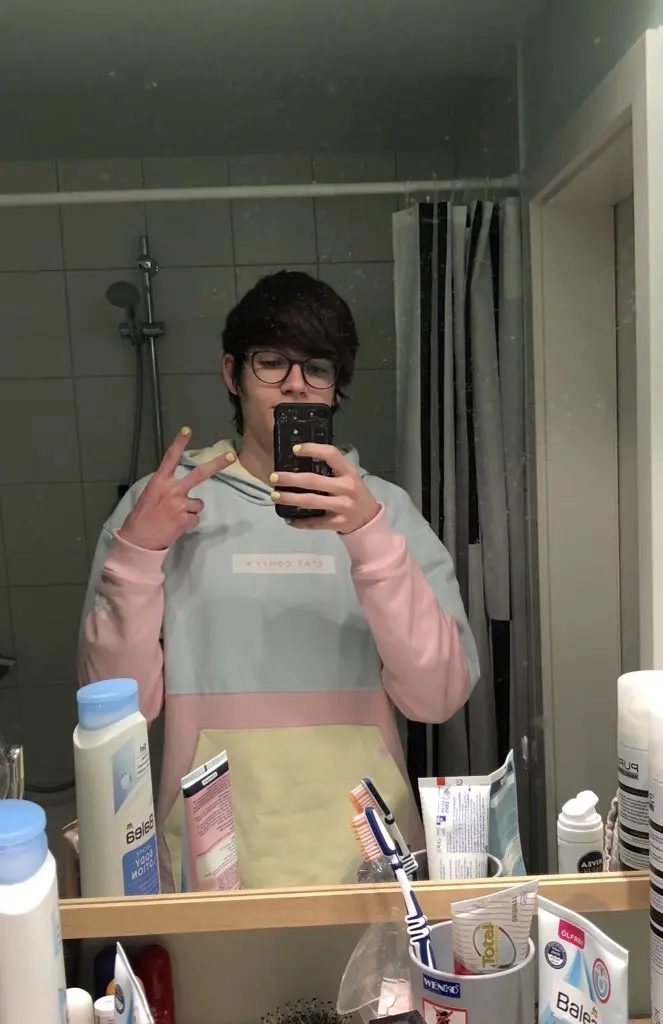 Photo of a person taking a mirror selfie in a bathroom. They have short dark hair, wear glasses, and a white and pink hoodie. They hold a black phone and make a peace sign with their free hand. The bathroom counter displays various toiletries including Balea products and toothbrushes in holders. White tile walls and a shower area are visible in the background.