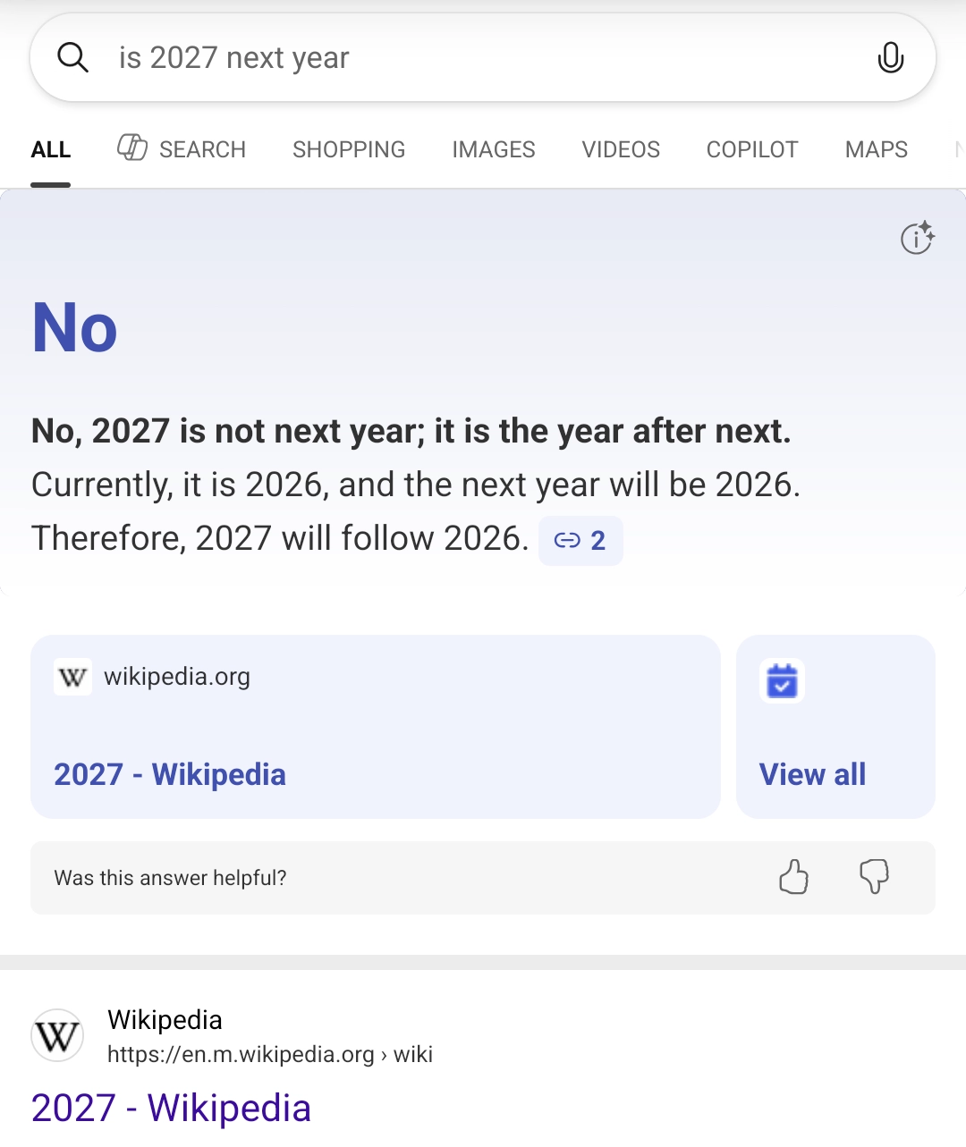 search results page with a light gray and white background. search query "is 2027 next year" is displayed in the search bar at the top with a magnifying glass icon on the left and a microphone icon on the right. The main answer box shows a large blue "No" at the top, followed by black text explaining: "No, 2027 is not next year; it is the year after next. Currently, it is 2026, and the next year will be 2026. Therefore, 2027 will follow 2026." with a blue link labeled "2". Below this are two light purple/blue rounded rectangular boxes - one containing the Wikipedia logo and text "wikipedia.org" with a purple link to "2027 - Wikipedia", and another box with a blue calendar icon and "View all" text. At the bottom is a feedback section asking "Was this answer helpful?" with thumbs up and thumbs down icons