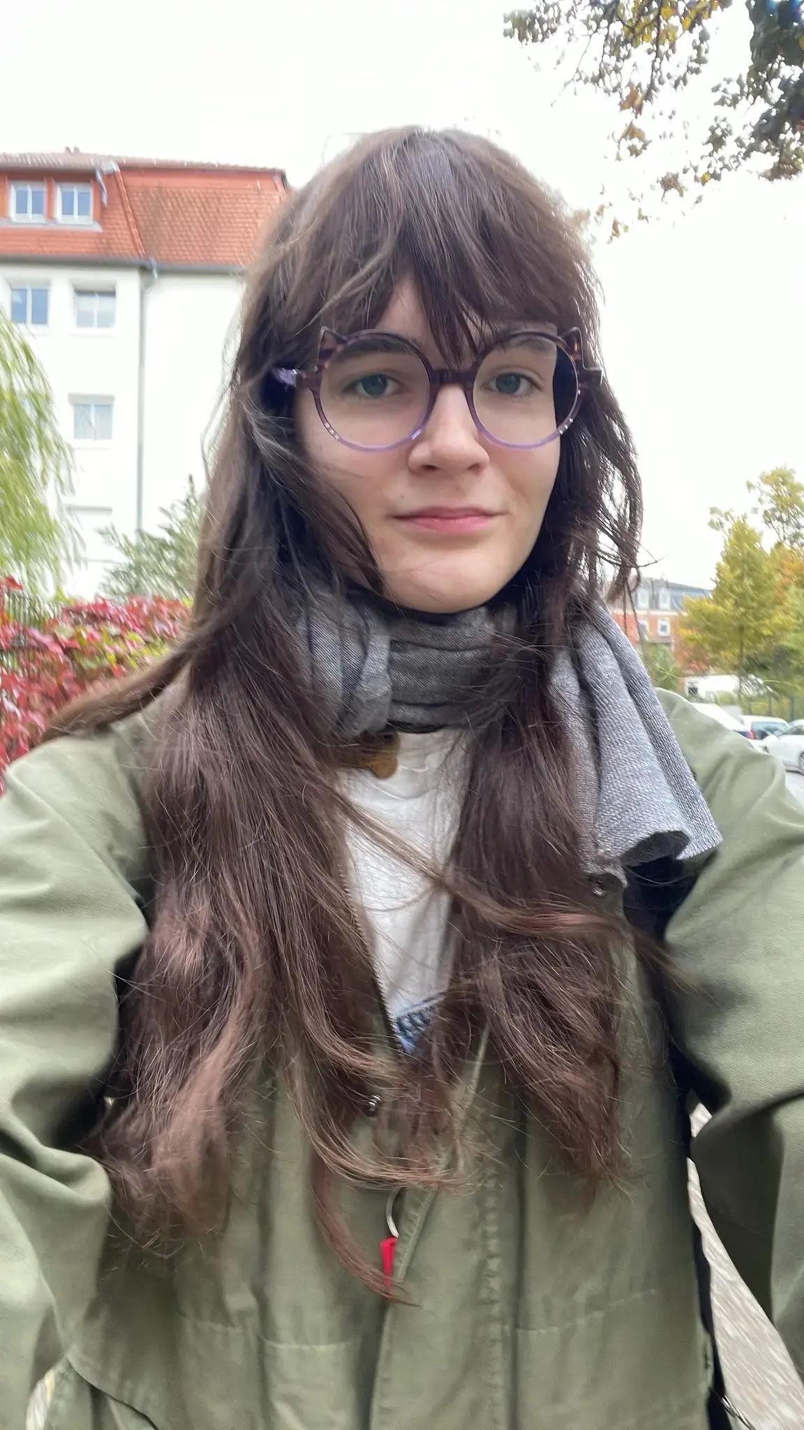 Photo of a person with long brown hair and purple-rimmed cat-eared glasses taking a selfie outdoors. They are wearing a gray striped scarf over a white shirt and a green jacket. Behind them is a white residential building with an orange-red tiled roof and blue windows, along with green and reddish autumn foliage. The setting is a residential neighborhood on an overcast day.