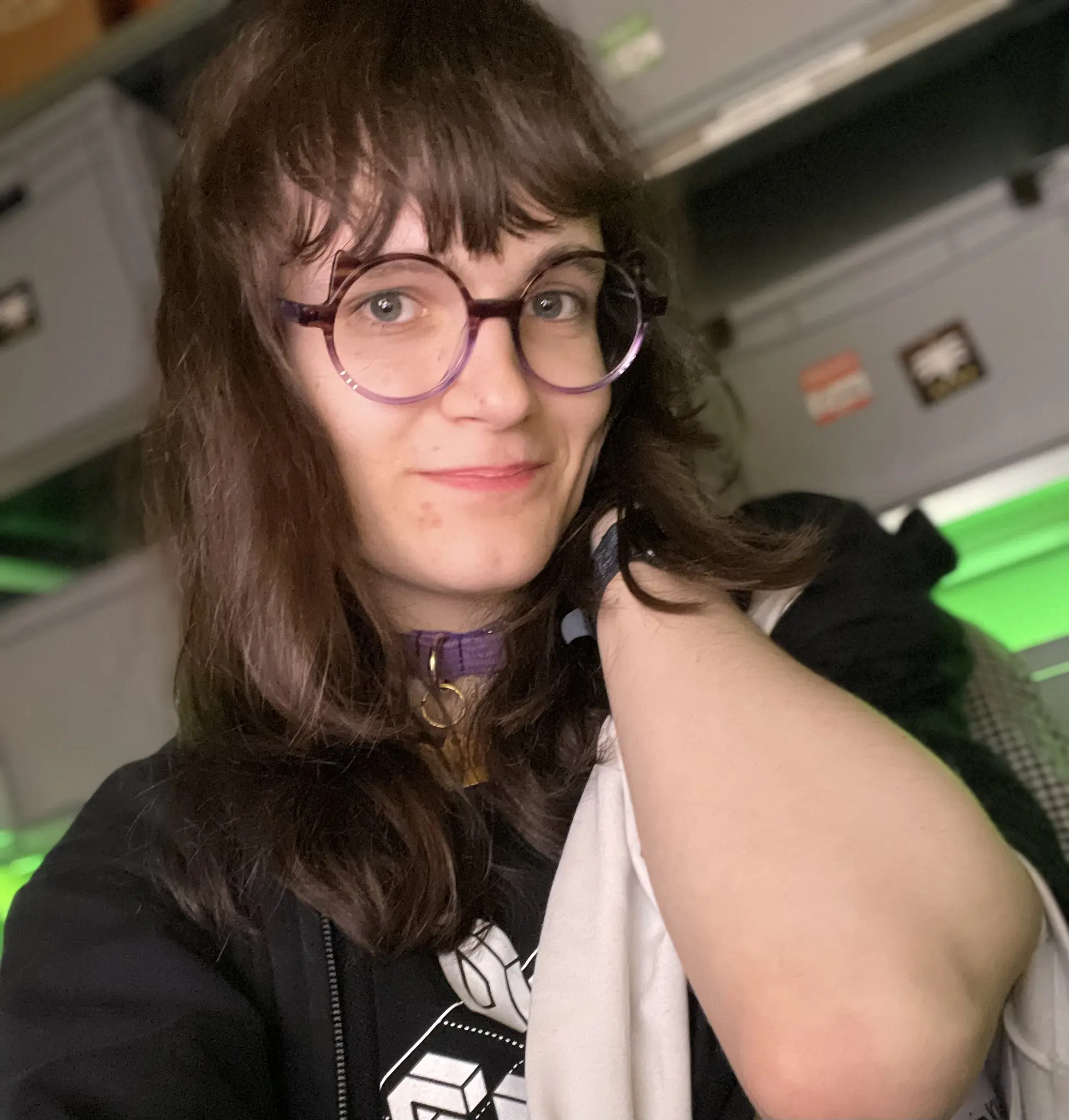 Photo. Person with shoulder length, slightly messy brown hair, a purple collar with gold bone charm and purple round glasses smiles at the camera with wide eyes. They wear a black hoodie over a black and white shirt, with one shoulder exposed showing white fabric underneath.