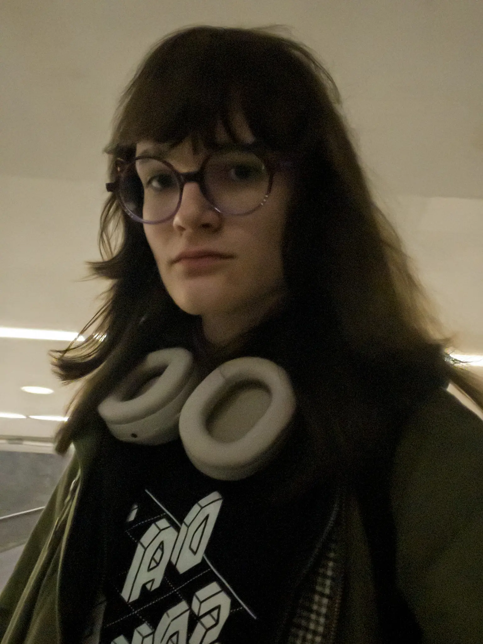 Photo taken from a low angle in dim lighting. Person with long brown hair and round glasses wears white over-ear headphones around their neck and a dark shirt with white 3d text and a green jacket which they are wearing unzipped. They are in a subway station with white light and a white ceiling